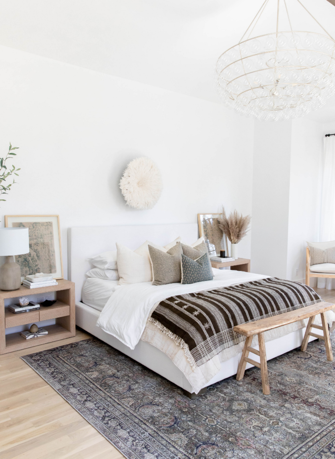 Large bedroom with king bed and night stands, brown throw blanket with cxream bedding, large gray designer rug with cream African juju  hat above the bed.