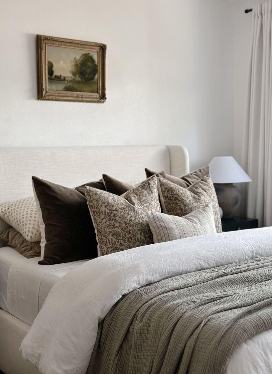 king bed with decorative neutral pillows with cream bedding, brown throw and small vintage art about bed.