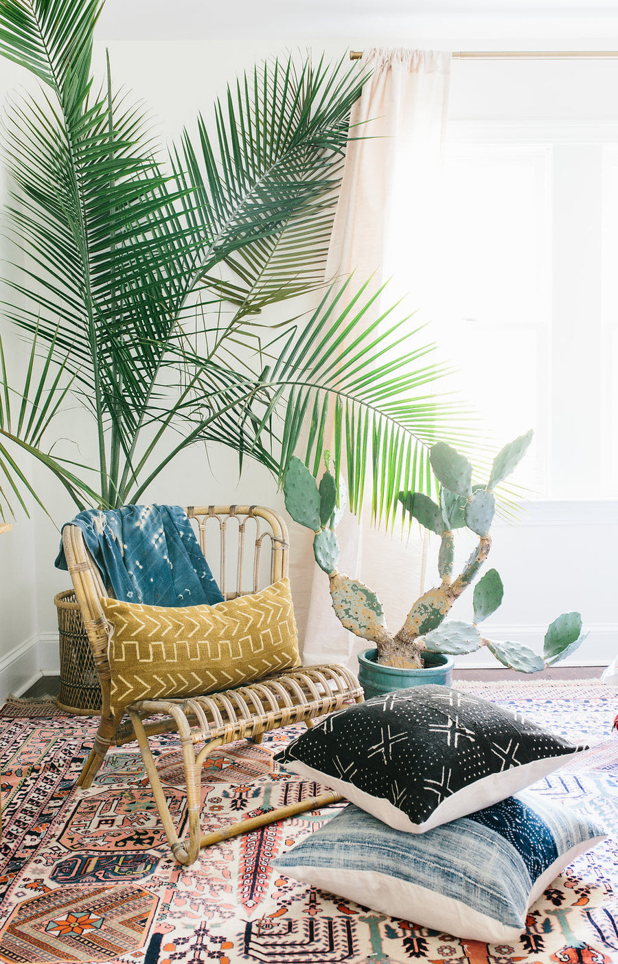 rattan chair with mustrad mudcloth pillow and large palm tree behind with two stacked mudcloth pillows on the floor and vintage multicolred rug