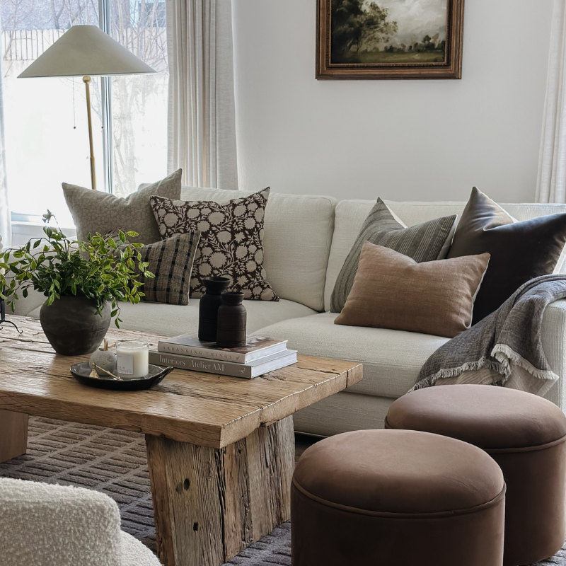 A styled living room scene featuring a cream-colored sofa decorated with multiple textured throw pillows, a rustic wooden coffee table, two round brown ottomans, a potted green plant, and a lamp in the background.