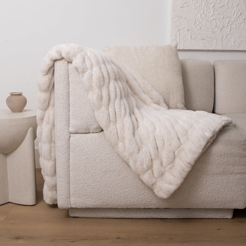 A chunky knit throw blanket in a warm cream or off-white color draped over a light beige textured sofa in a minimalist living room setting.