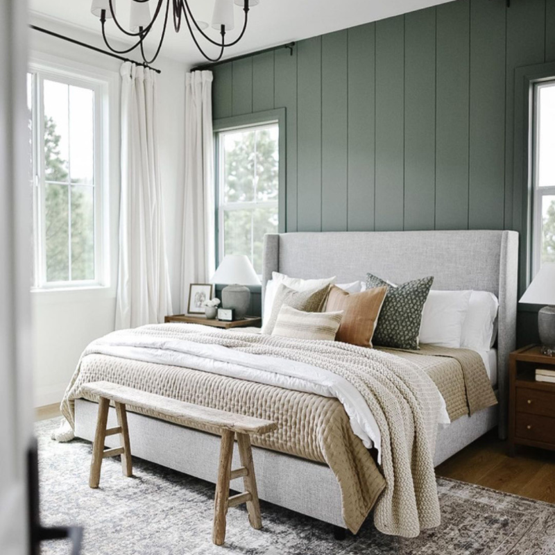 A styled bedroom featuring a large upholstered bed with a high back, dressed in beige and white textured bedding and decorative pillows. The room has a prominent dark green paneled wall, white curtains, wooden nightstands, and a rustic wooden bench at the foot of the bed.